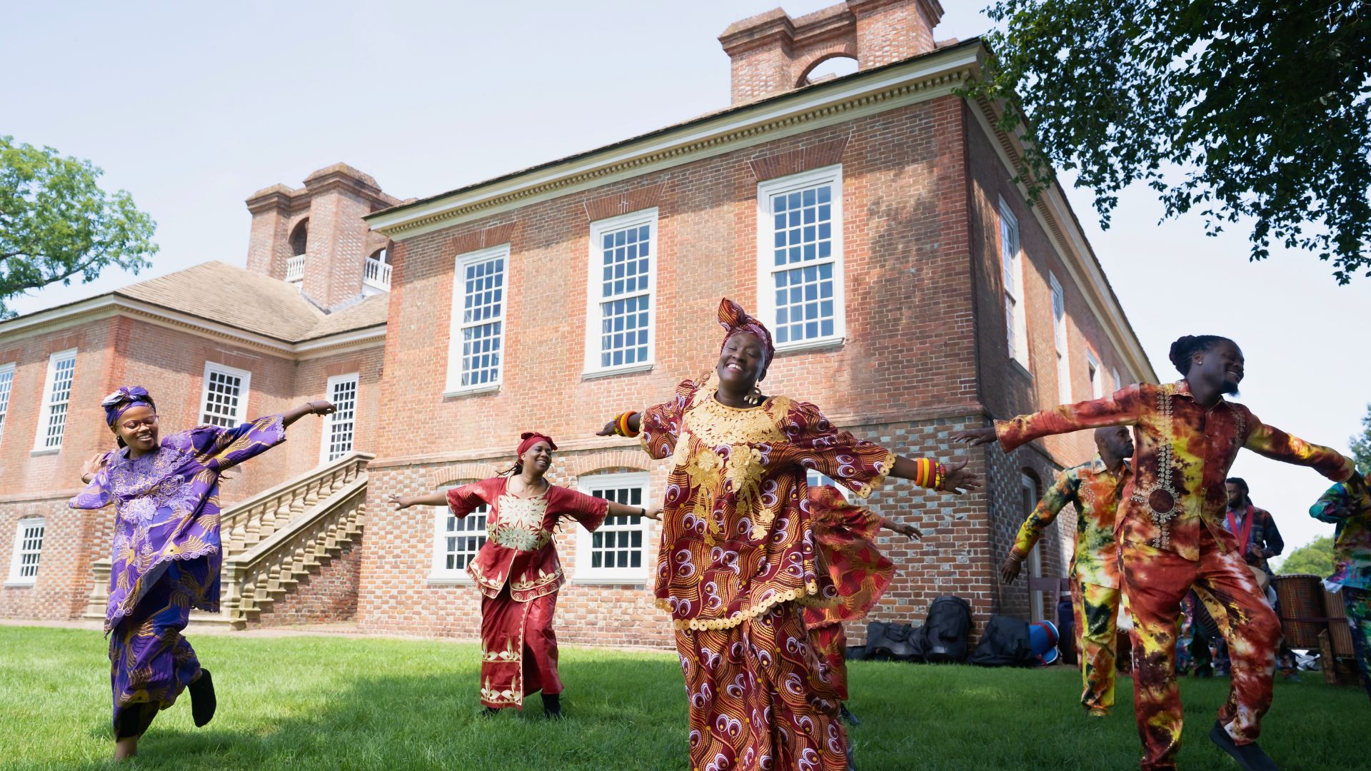 Group of people in vibrant African attire dancing on a lawn in front of a brick campus building (outdoor cultural performance).