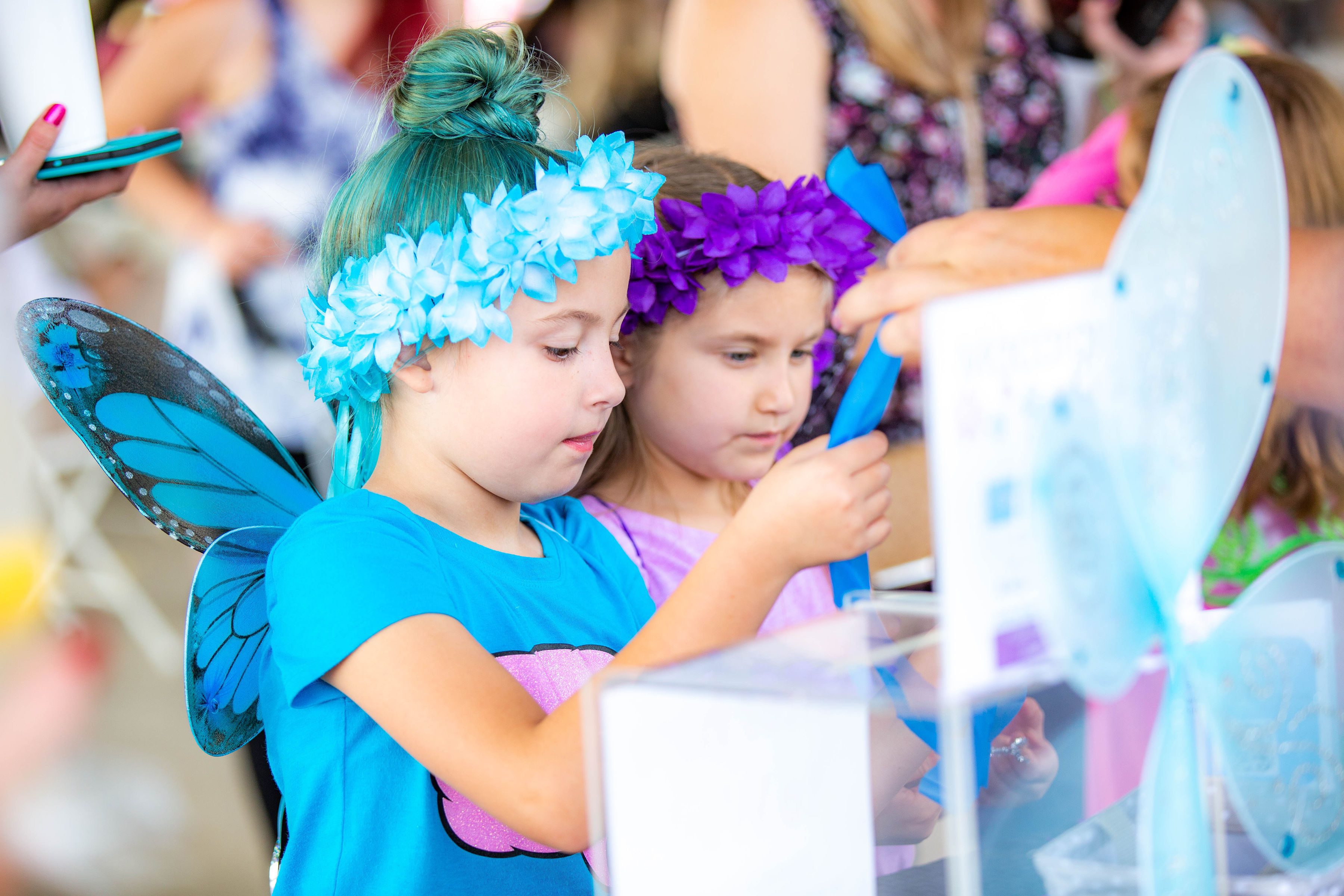 Two young girls in butterfly costumes with blue wings and flower headbands crafting at a booth.