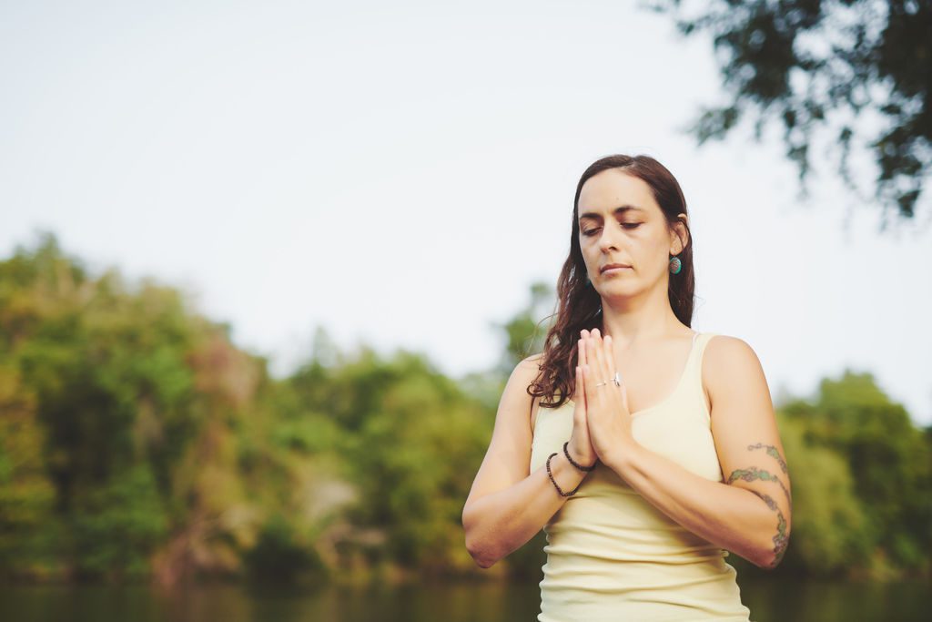 Woman in a pale yellow tank top meditating outdoors, hands pressed in prayer by a lakeside.